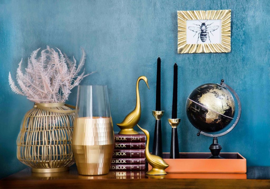 assorted-gold-plated-table-figurines-1099816 Stylish home decor arrangement including a globe, books, candles, and vases on a blue backdrop.